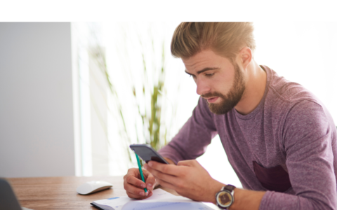 Person taking notes in a book while looking at his phone